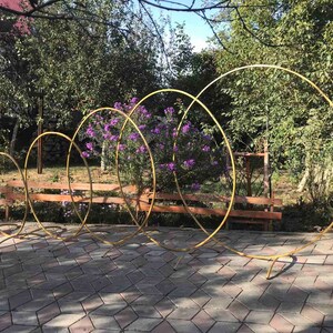 May include: Five gold metal hoops of varying sizes stand upright on a gray brick patio. The hoops are arranged in a staggered formation, with the largest hoop in the back and the smallest hoop in the front. The hoops are all made of a single piece of metal and have a smooth, polished finish.