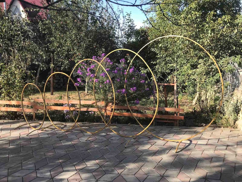 May include: Five gold metal hoops of varying sizes stand upright on a gray brick patio. The hoops are arranged in a staggered formation, with the largest hoop in the back and the smallest hoop in the front. The hoops are all made of a single piece of metal and have a smooth, polished finish.
