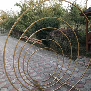May include: A set of four gold metal hoops, arranged in a concentric circle pattern, creating a decorative archway. The hoops are supported by a sturdy metal base.