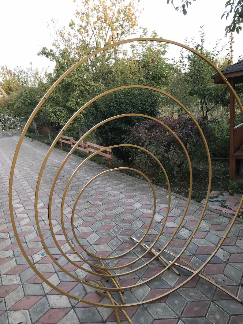 May include: A set of four gold metal hoops, arranged in a concentric circle pattern, creating a decorative archway. The hoops are supported by a sturdy metal base.