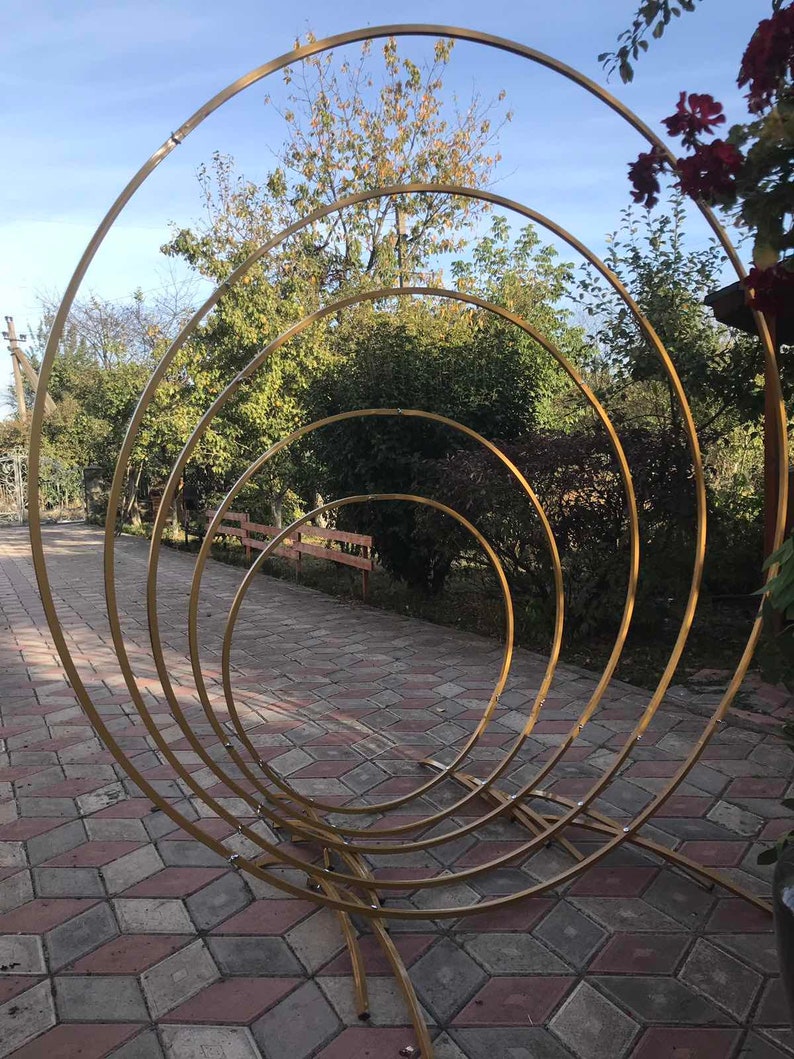 May include: A gold metal archway made of four concentric circles. The archway is standing on a brick patio.