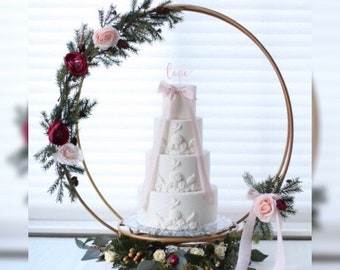 May include: A white four-tier wedding cake with pink ribbon and floral details sits on a gold cake stand with a gold hoop decorated with greenery and pink and burgundy flowers. The hoop has the word "love" written on it in pink.