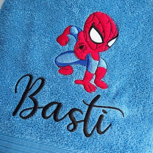 Könnte beinhalten: Blaues Badetuch mit gesticktem Spider-Man-Motiv in Rot, Blau und Weiß. Der Name "Basti" ist in schwarzer Schrift gestickt. Das Handtuch hat eine strukturierte Oberfläche und einen hellblauen Rand, ideal für Kinder.