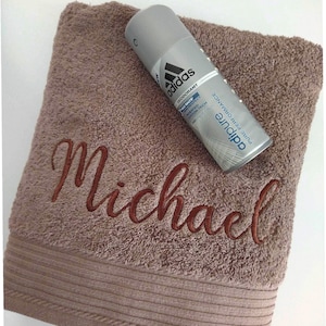 Könnte beinhalten: Ein braunes Handtuch mit dem Namen "Michael" in dunklerbrauner Schrift gestickt. Ein silbernes Adidas Deodorant-Spray liegt auf dem Handtuch. Das Handtuch hat eine strukturierte Oberfläche und eine dekorative Bordüre.