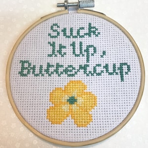 May include: A cross-stitch embroidery hoop with the phrase "Suck It Up, Buttercup" in green thread. A yellow flower with a green center is stitched below the text. The embroidery is set in a wooden hoop.