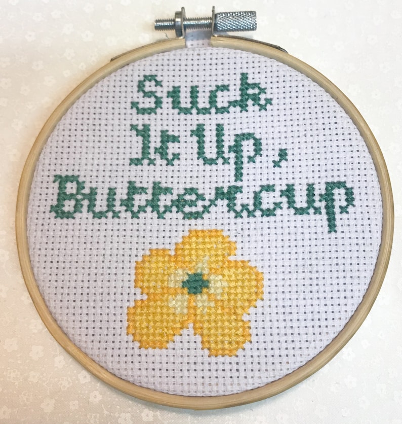 May include: A cross-stitch embroidery hoop with the phrase "Suck It Up, Buttercup" in green thread. A yellow flower with a green center is stitched below the text. The embroidery is set in a wooden hoop.