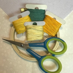 May include: A craft kit with embroidery floss in shades of yellow and green, a wooden hoop, and blue and green handled scissors. A piece of white cross-stitch fabric is in the background.