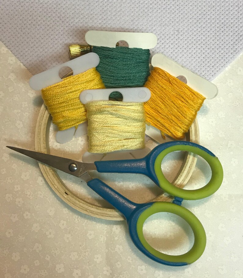 May include: A craft kit with embroidery floss in shades of yellow and green, a wooden hoop, and blue and green handled scissors. A piece of white cross-stitch fabric is in the background.