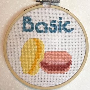 Puede incluir: Un aro de bordado blanco con la palabra "Basic" bordada en hilo azul. Dentro del aro hay dos macarons bordados, uno amarillo y otro rosa.