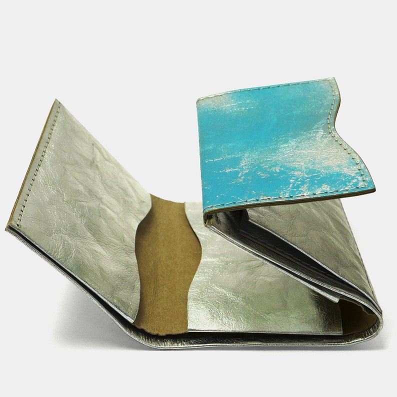 May include: A silver leather wallet with a blue leather flap. The wallet is open and the inside is visible. The wallet has multiple compartments for cards and cash.