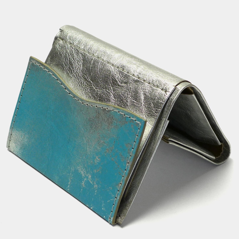 May include: A silver leather wallet with a turquoise blue interior. The wallet is folded and the interior is visible.