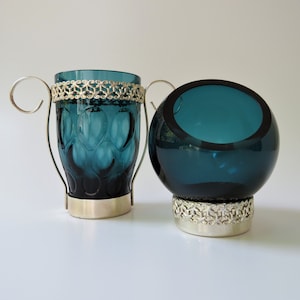 Può includere: Due portacandele in vetro blu turchese con basi in metallo argentato e decorazioni decorative. I portacandele sono su una superficie bianca.