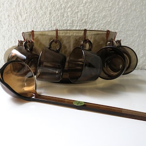 Può includere: Una ciotola per punch in vetro marrone con sei tazze abbinate e una paletta in plastica marrone. La ciotola ha un design decorativo e le tazze hanno manici.