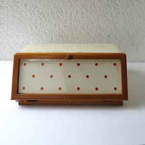 May include: A vintage wooden bread box with a cream-colored top and a glass door. The door features a grid of red polka dots. The box has a wooden frame and a small wooden knob for opening.