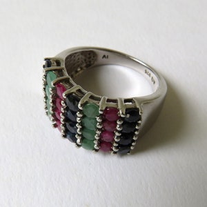 Puede incluir: Un anillo de plata con una banda de piedras preciosas de esmeralda, rubí y zafiro. El anillo está marcado con "Talla 6" y "A1" en el interior.