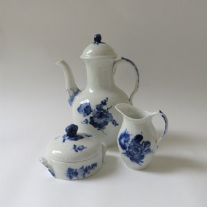 Può includere: Un servizio da tè in porcellana bianca con disegni floreali blu. Il set comprende una teiera, una lattiera e una zuccheriera con coperchio. La teiera ha un manico e un beccuccio curvi. La lattiera ha un manico curvo.