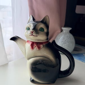 Op de afbeelding: Een keramische theepot in de vorm van een kat, zwart en crème. De theepot heeft groene ogen, een rode strik en een gebogen handvat. De tuit komt uit de poot van de kat. Een decoratief item.