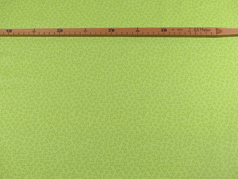 以下が含まれることがあります： 小さな手書きの三角形が繰り返しパターンになっている緑色の生地。生地の上に、インチとセンチの目盛りがついた木製の定規が置かれている。