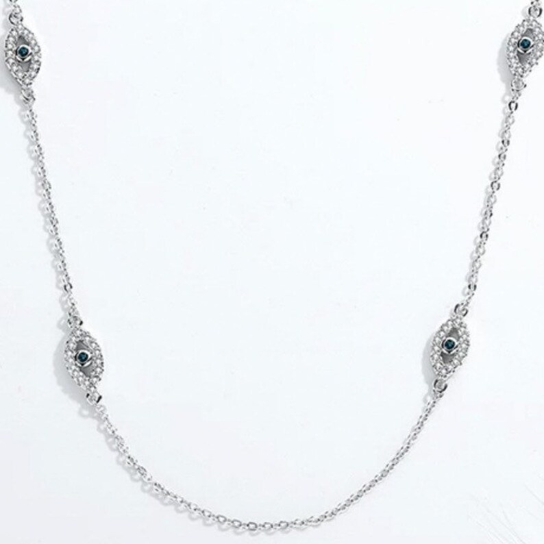 May include: A silver-toned chain necklace featuring multiple eye-shaped charms. Each charm is encrusted with small clear stones and centers on a single dark blue stone. The delicate metal chain connects these symmetrical motifs in a repeating pattern.