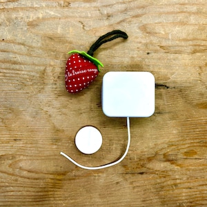 Puede incluir: Una caja de música blanca con una cuerda de tracción y un encanto de tracción en forma de fresa roja y blanca con el texto "la fraise rouge".