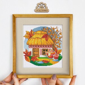 Puede incluir: Imagen de punto de cruz de una cabaña con techo de paja, enmarcada en madera. La escena representa un paisaje otoñal con un árbol, hojas cayendo y un juego de té. La obra de arte está sostenida por manos.