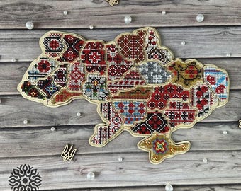 May include: A wooden map of Ukraine with a colorful embroidered design. The map is made up of many small squares, each with a different pattern. The map is surrounded by white pearls and a wooden trident.