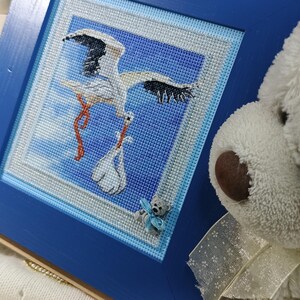 May include: A framed cross-stitch artwork featuring a stork carrying a baby in a bundle. The artwork is set against a blue sky background and framed in a blue wooden frame. A small teddy bear is attached to the frame. A white teddy bear is in the background.