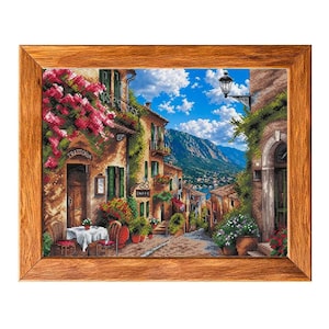 May include: A framed cross-stitch embroidery depicting a Mediterranean street scene with stone buildings, pink bougainvillea, a small table with chairs, and the text Trattoria and Caffe, set against a backdrop of mountains and a blue sky.