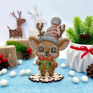 Könnte beinhalten: Weihnachtsdekoration aus Holz, die ein Rentier mit Hut und Geschenk zeigt. Das Rentier ist braun mit einem rot-grünen Schal und einer Schneeflockenbasis. Weitere dekorative Rentiere und festliche Akzente befinden sich im Hintergrund.