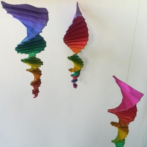 Könnte beinhalten: Drei hängende, abstrakte Skulpturen in Regenbogenfarben. Jede Skulptur hat einen gefalteten, fächerartigen oberen Teil und einen spiralförmigen, konischen unteren Teil. Die Farben wechseln von Lila zu Rot, dann zu Gelb und Grün. Die Skulpturen sind vor einer weißen Wand aufgehängt.