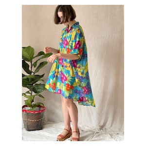 May include: A vibrant, short-sleeved dress with a colorful floral pattern in shades of blue, pink, yellow, and red. The dress has a collared neckline and a slightly asymmetrical hemline. The model is wearing brown wedge sandals.