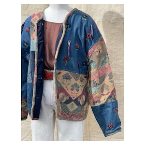 Peut inclure: Veste en jean patchwork avec broderies florales et panneaux de tissu à motifs. La veste présente un mélange de denim bleu, de tissu matelassé bleu marine et de sections de type tapisserie avec des motifs roses, beiges et verts. Coupe décontractée, bordure marron.
