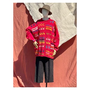 May include: A vibrant red, oversized sweater with colourful abstract patterns. The sweater has a crew neck and long sleeves. The mannequin is also wearing black culottes and a brown hat. The background is a mix of red and white fabric.