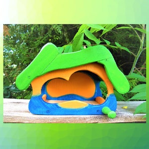 Op de afbeelding: Een houten vogelvoederhuisje met een felgroen dak, een oranje achterwand aan de binnenkant en een blauwe basis. De structuur heeft een brede, open ingang aan de voorzijde en een klein groen pinnetje dat aan de rechterkant uit de blauwe basis steekt.