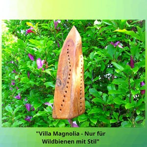 Könnte beinhalten: Ein bienenfreundliches Holzhaus mit dreieckiger Form und zahlreichen Bohrlöchern. Das Bienenhaus steht vor einem Hintergrund aus grünem Laub und lila Blüten. Der Text unten lautet "Villa Magnolia - Nur für Wildbienen mit Stil."