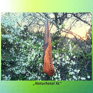 Può includere: Una scultura in legno realizzata a mano con finitura marrone scuro e una serie di fori praticati. La scultura è sospesa su uno sfondo di alberi in fiore bianchi e un tramonto. Il testo "Naturhotel XL" è in basso.