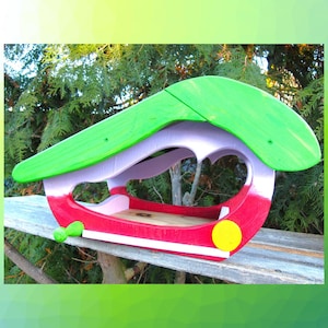 May include: A wooden bird feeder featuring a bright green curved roof, a white middle section, and a red base. The structure includes a small yellow circular accent on the side and a natural wood interior floor for bird seed placement.