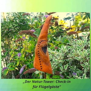 Puede incluir: Una escultura de madera naranja con una serie de pequeños agujeros y una abertura más grande, colocada en un jardín. El jardín presenta follaje verde y blanco, flores moradas y flores amarillas. El texto en la parte inferior dice "Der Natur-Tower: Check-in für Flügelgäste."
