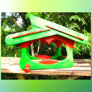 May include: A wooden bird feeder painted in bright green and red. The structure features a slanted roof, open arched sides for access, and a small red cylindrical perch extending from the front base. The design is symmetrical with smooth edges.