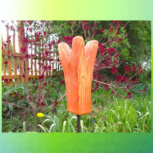 Può includere: Un paletto da giardino a forma di tulipano in legno con finitura arancione si trova in un giardino erboso. Il paletto ha una base quadrata e una forma di tulipano stilizzata con tre petali. Lo sfondo comprende una staccionata in legno, cespugli con foglie rosse ed erba verde.