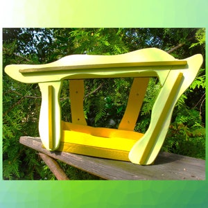 May include: A wooden bird feeder featuring a bright yellow base and light lime green side panels with curved, organic edges. The structure has an open design with four vertical supports connecting the base to a wide, overhanging roof.