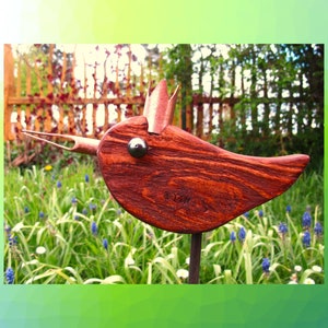 Könnte beinhalten: Eine handgefertigte Holzskulptur eines Vogels mit dunkelrotbraunem Finish. Der Vogel hat einen langen Schnabel, ein Metallauge und eine Metallkrone. Er ist auf einer Metallstange montiert und in einem Garten mit grünem Gras und blauen Blumen aufgestellt.