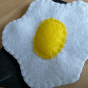 May include: A white felt fried egg with a yellow felt yolk, sewn with a decorative stitch around the edges.