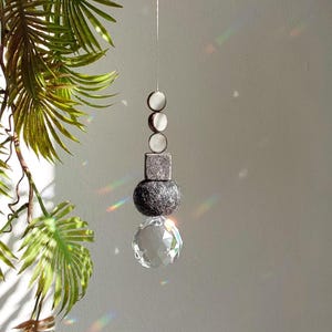 Puede incluir: Un atrapasol de cristal con una bola de fieltro gris, un cuadrado gris y tres cuentas redondas blancas colgando de una cuerda.