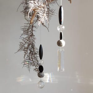 Könnte beinhalten: Hängendes Mobile mit einem natürlichen Ast und getrocknetem Laub. Das Mobile hat zwei Stränge mit schwarzen ovalen Perlen, klaren Glasperlen, grauen Steinperlen und einem klaren rechteckigen Kristall.