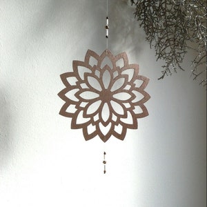Op de afbeelding: Een decoratieve, hangende mobiel met een bloemvormig ontwerp, uitgesneden uit metaalachtig bruin materiaal. De mobiel hangt aan een dun touwtje met kleine kralen en een kristallen hanger. De achtergrond is een witte muur.