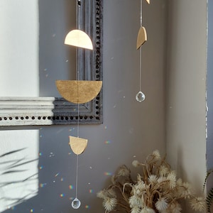 Peut inclure: Un mobile géométrique doré avec des prismes de cristal suspendus. Le mobile est en bois et a un design moderne. Il est suspendu dans un coin d'une pièce avec un mur blanc et un cadre photo blanc.