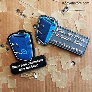 Peut inclure: Deux patchs brodés gris et bleus avec une image de dessin animé d'un appareil avec des boutons et un cercle. Le patch du haut porte le texte "Mike...No Shoot...No Shoot...Mike...but check out the Splits". Le patch du bas porte le texte "Game plan disappears after the beep".