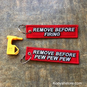 May include: Two red keychains with white text. The top keychain says "REMOVE BEFORE FIRING". The bottom keychain says "REMOVE BEFORE PEW PEW PEW". A yellow plastic key chain puller is also included.