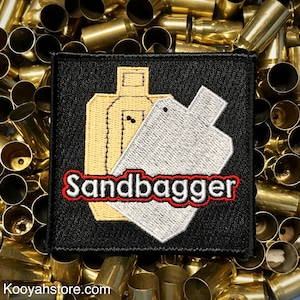 May include: Black embroidered patch with a yellow and white target silhouette and the text "Sandbagger" in red with a white outline.